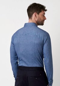 Blauwe langlopende blouse van een gestructureerde stof, met een klassieke kraag en een op maat gemaakte achterkant, gecombineerd met donkere broeken.