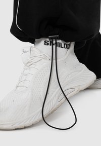 Weißer Sportschuh mit strukturiertem Obermaterial, schwarzen Schnürsenkeln und ausgeprägter Gummisohle. Schwarzes Elastikband vom taillierten Hosenbein am Knöchel gesichert.