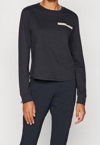 Black long-sleeve shirt featuring a chest pocket with a beige detail. Made of soft fabric with a relaxed fit and stitched hem.