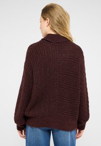 Burgundijas adīts džemperis ar augstu apkakli, ar teksturētām rakstiem un ribotām piedurknēm, apvienots ar gaiši zilām džinsu biksēm.