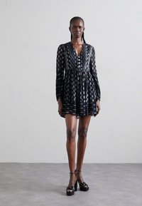 Schwarzes Kleid mit langen Ärmeln, tiefem V-Ausschnitt und gemustertem Stoff. Mit einem gerafften Bund und knielangem Saum, kombiniert mit Plateauschuhen.