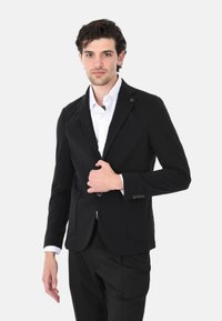 Blazer nero con revers, chiusura a un bottone, tasche frontali e tessuto texturizzato. Indossato sopra una camicia bianca.