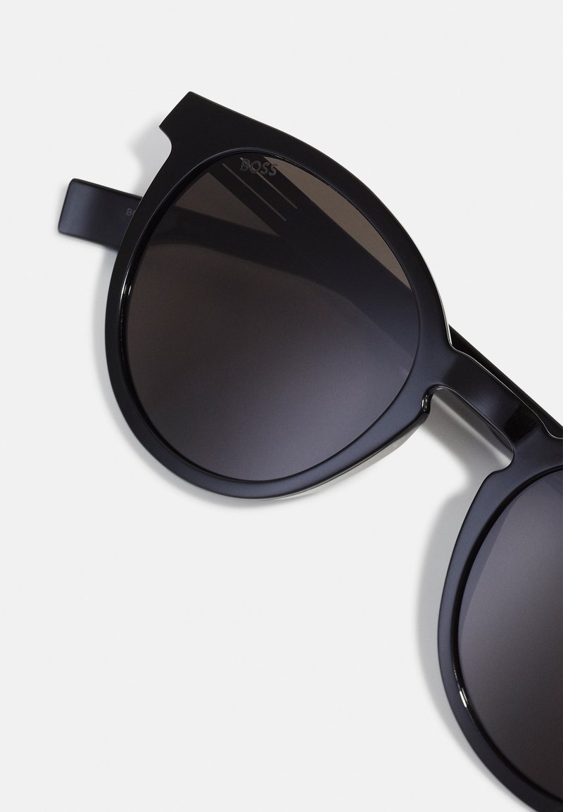 Occhiali da sole neri con una montatura spessa e angolare, lenti scure a gradiente e un logo chiaro sul lato superiore sinistro della montatura.