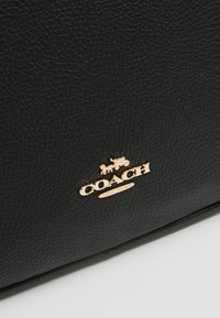 Black textured leather surface with a gold Coach New York logo featuring a horse-drawn carriage emblem.