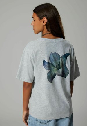Camiseta de algodón gris con mangas cortas, que presenta un gran gráfico detallado de una flor azul en la parte posterior. Textura suave, ajuste casual.