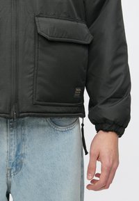 Casaco acolchoado preto com punhos elásticos, bolso frontal e etiqueta com logótipo. Textura suave. Usado com jeans azul claro.