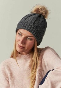 Bonnet en tricot gris avec une texture côtelée, ornée d'un pompon fluffy couleur brun clair sur le dessus. L'arrière-plan est neutre.