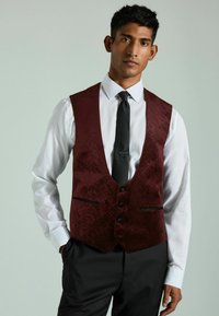Chaleco burdeos con estampado, con escote en V, tres botones y detalles en negro, usado sobre una camisa blanca y una corbata negra. Corte entallado.