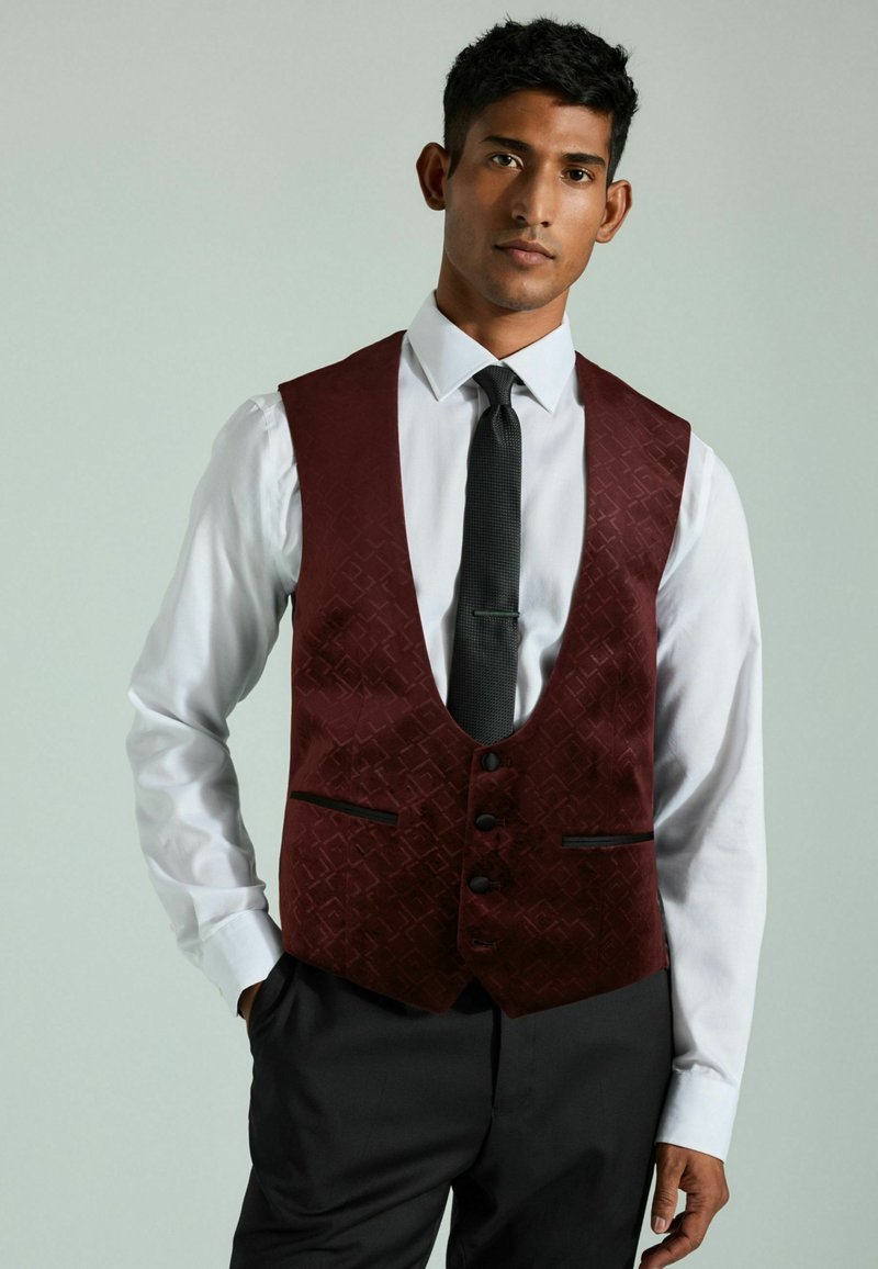 Chaleco burdeos con estampado, con escote en V, tres botones y detalles en negro, usado sobre una camisa blanca y una corbata negra. Corte entallado.