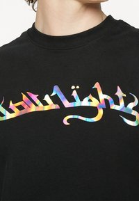 Nahaufnahme eines schwarzen Shirts mit mehrfarbigem gotischem Text über der Brust, getragen von einer Person mit sichtbarem Hals und Kiefer.