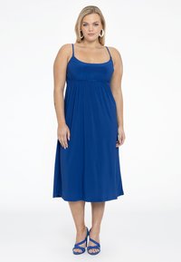 Blå ærmeløs maxikjole med en samlet talje, lavet af blødt stof. Kombineret med blå strop-sandaler og store hvide hoop øreringe.