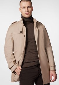 Beżowy płaszcz trench z wysokim kołnierzem, guzikami z przodu i naramiennikami. Nosi się go na ciemnobrązowy golf i czarne spodnie.