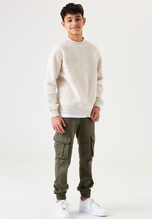 Fleece sweatshirt in crèmekleur met een textuur en de tekst "NIET GEMIDDELD"; olijfgroene cargobroek met elastische boorden; witte sneakers.