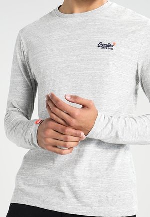 Mężczyzna w jasno szarej koszuli z długim rękawem, z małym czarno-pomarańczowym logo na piersi, trzymający jedną dłoń drugą przed sobą.