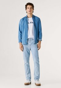 Junger Mann, der hellblaue Jeans, ein weißes T-Shirt mit Aufdruck, ein aufgeknöpftes blaues Jeanshemd und weiße Sneakers trägt, vor einfarbigem Hintergrund steht.