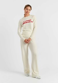 Crèmekleurige gebreide trui met rode "J'adore Christmas" tekst, gecombineerd met bijpassende crème kleurige flared broek en lichtgekleurde sneakers.