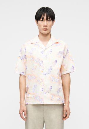 Man wearing short-sleeve button-up shirt with pastel purple and orange butterfly print, paired with light beige pants, standing still.