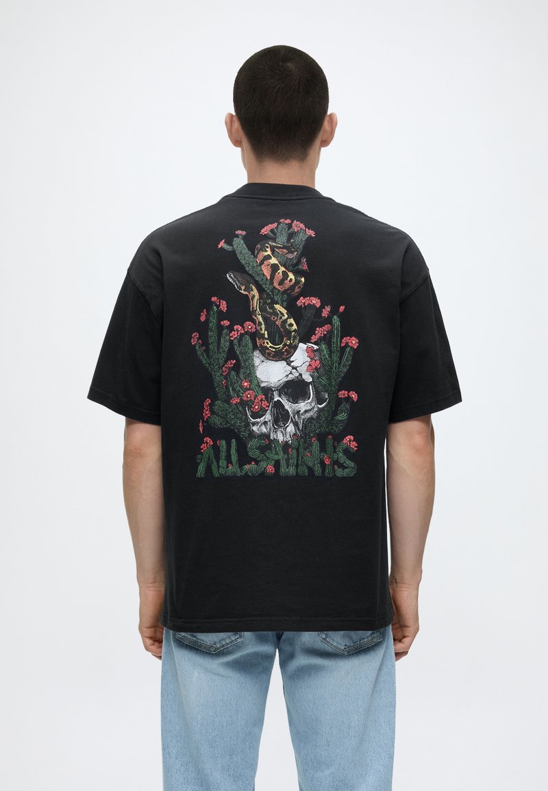 Mężczyzna noszący czarny t-shirt z grafiką przedstawiającą węża owiniętego wokół czaszki i kaktusów otoczonych różowymi kwiatami, wraz z napisem „AllSaints”.