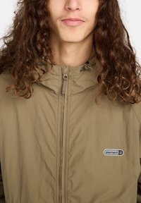 Persoon met lang, krullend haar in een lichtbruine zip-up jas met een Element-logo op de borst, tegen een effen achtergrond.
