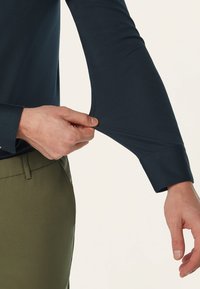 Marineblauwe longsleeve shirt met een nauwsluitend ontwerp, met een subtiel textuurstof. Een hand trekt aan de mouw, waardoor een gladde afwerking zichtbaar wordt.