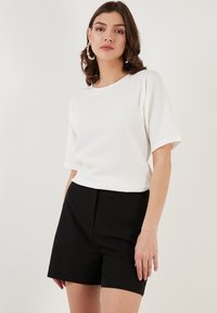 Witte textuur korte mouwtop met een ronde halslijn, gecombineerd met hoge taille zwarte shorts. Model draagt grote hoepeloorbellen.