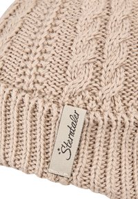 Beige knitted fabric with ribbed and braided patterns, featuring a rectangular beige label with cursive text sewn onto the material.