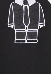 Camicia in tessuto nero con grafica ricamata di un completo con cravatta, contornata in bianco, che mostra un design di colletto e pantaloni.