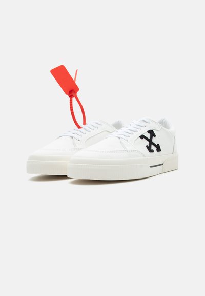 Λευκά sneakers τύπου low-top με μαύρο λογότυπο σταυρωμένου βέλους στο πλάι και κόκκινη πλαστική ζώνη φερμουάρ στο αριστερό παπούτσι.