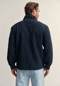 Tamsiai mėlynas flisinis megztinis su aukštu apykakliu, tekstūruota medžiaga ir ribinėmis rankogaliais; dėvimas su šviesiai mėlynomis džinsais, matomas iš nugaros.