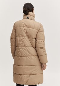 Vadderad beige lång kappa med en hög krage, med horisontella kviltade sektioner och en triangel-logotypdetalj på baksidan.