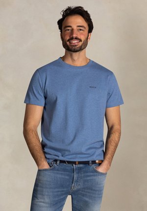 Glimlachende man met donker haar, gekleed in een lichtblauw shirt met korte mouwen en een blauwe jeans, handen in de zakken, tegen een effen achtergrond.