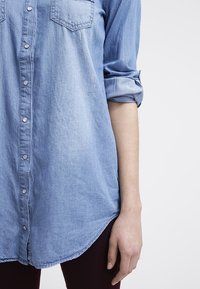 Ljust blå denimskjorta med knäppning framtill, två bröstfickor, uppvikta ärmar och en rundad fåll. Mjuk textur.