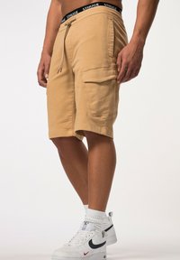 Shorts cargo beiges avec taille élastique et cordon de serrage, dotés de poches latérales et d'une coupe décontractée. Portés avec des baskets blanches Nike.