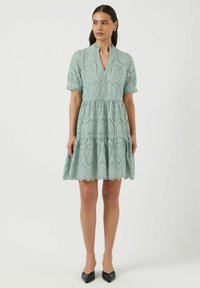 Svetlo zelena obleka s plastnim dizajnom, vzorcem z očesci, kratkimi napihnjenimi rokavi in ovratnikom s pokritimi robovi, ki se konča malo nad kolenom.