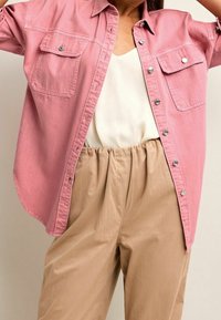 Camisa de mezclilla rosa con dos bolsillos en el pecho, detalles de botones plateados, llevada sobre una camiseta beige y pantalones de cintura elástica en taupe.