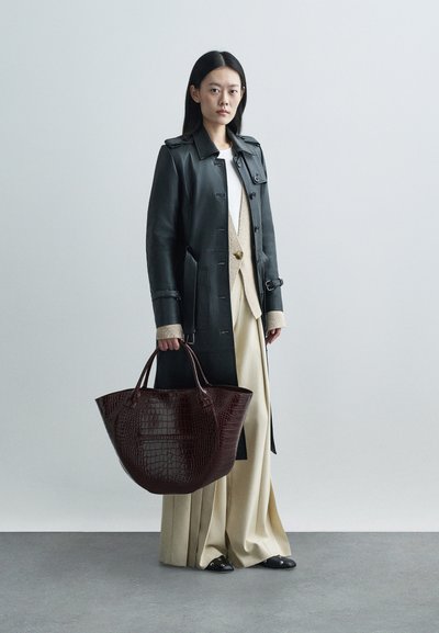 Μαύρο δερμάτινο παλτό με μεγάλα κουμπιά, συνδυασμένο με κρεμ πουλόβερ και φαρδιά μπεζ παντελόνια. Κρατώντας τσάντα tote σε μπορντώ με ανάγλυφο σχέδιο κροκοδείλου.