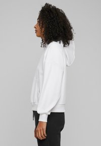 Weiße Kapuzenpullover mit einer Vordertasche und lockerer Passform. Verfügt über gerippte Bündchen und Saum, getragen mit schwarzen Hosen.
