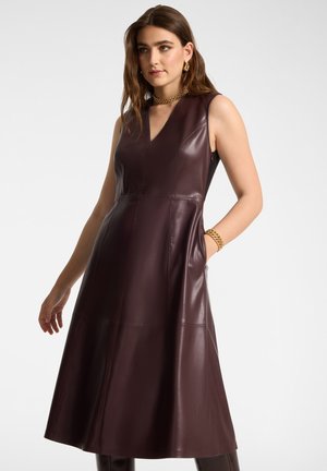 Femme portant une robe en cuir marron foncé sans manches en ligne A avec un col en V, accessoirisée d'un collier chaîne en or, d'un bracelet et de boucles d'oreilles, posant avec la main dans la poche.
