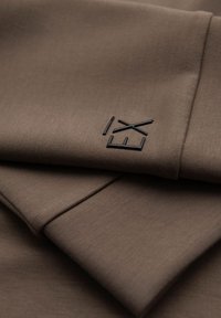 Tela marrón oscura con una textura suave, con un logotipo negro en relieve que dice "EX" en un lado. El diseño es simple y minimalista.