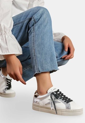 Une personne en jean bleu est accroupie, portant des baskets blanches avec des lacets noirs, des détails en paillettes argentées et des accents de imprimé léopard.