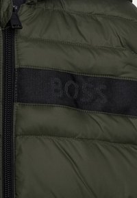 Close-up of olive green quilted jacket with black horizontal stripe and vertical black zipper on left side.