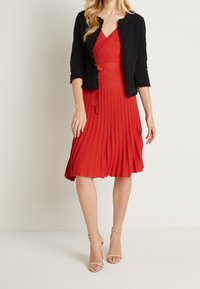 Červené plisované šaty s překříženým designem, v kombinaci s černým zkráceným kardiganem s volánky. Outfit doplňují béžové lodičky.