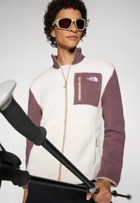 Fleece jack met een witte body en mauve mouwen en borstzak. Rits sluiting en logo detail. Accessoires zijn trekkingstokken.