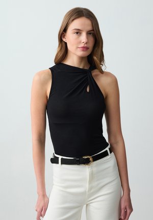 Top a fascia nero a coste con un dettaglio frontale torcato e un'apertura a chiave, abbinato a pantaloni bianchi a vita alta e una cintura nera con accessori dorati.