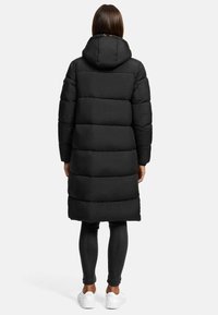 Svart kapuschong puffjacka med en lång, vadderad design. Har en texturerad yta, stora paneler och en dragkedja fram. Burits över mörka leggings och vita skor.