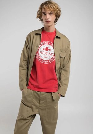 Camisa caqui de botones sobre una camiseta gráfica roja con texto blanco, combinada con pantalones caqui holgados que tienen bolsillos laterales.