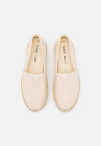 Beżowe płócienne buty typu slip-on z zaprojektowanym w kształcie litery V przeszyciem oraz podeszwą z juty. Wewnątrz buta widoczna metka marki. Płaska, okrągła czubek.