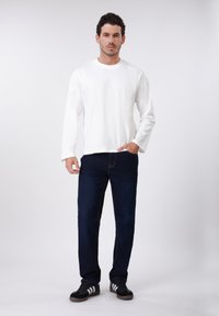 Λευκή μπλούζα με μακριά μανίκια, σκούρο μπλε τζιν, αθλητικά παπούτσια ασπρόμαυρα, σε στάση όρθια μπροστά σε απλό φόντο.