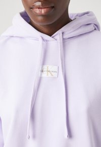Hoodie lilla con cordini, dotato di un'etichetta che recita "Calvin Klein Jeans" in lettere dorate. Tessuto morbido con una texture liscia.