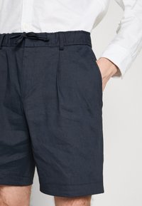 Tamsiai mėlynos šortai, pagaminti iš lengvo audinio, su elastingu juosmeniu ir raišteliu, taip pat turi šonines kišenes, skirtos patogumui.